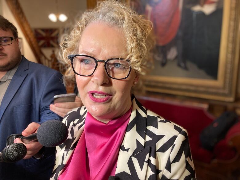 A woman with blond hair speaks into a microphone in a group of reporters.