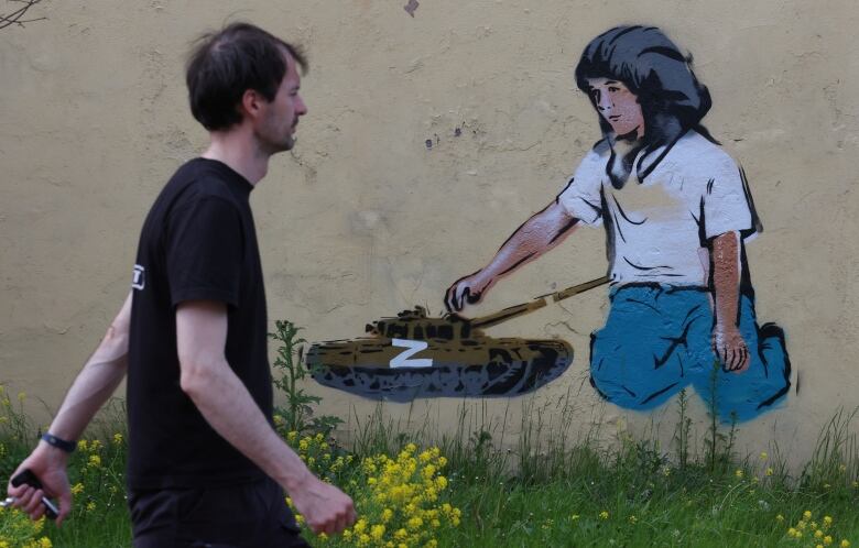 A man in a black T-shirt walks past graffiti depicting a tank with the Z symbol.