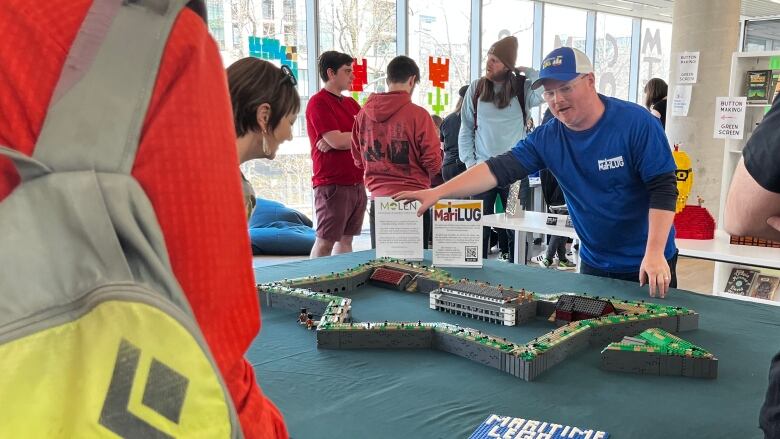 A man gestures towards a Lego replica of Citadel Hill as a small crowd gathers around the table. 