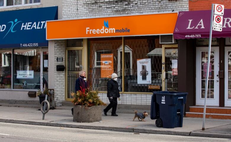 Two people walk past a Freedom Mobile store with orange hoardings.