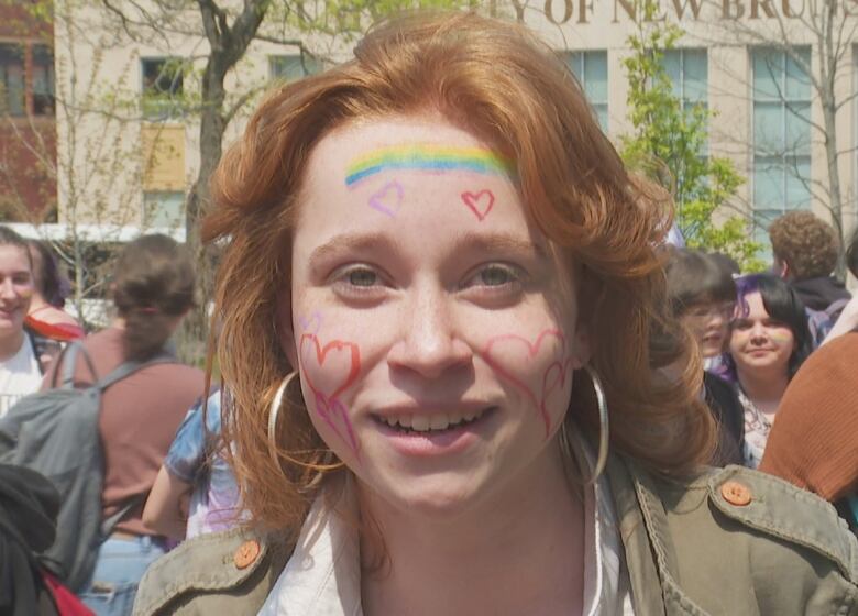 A person with red hearts drawn on their cheeks and a rainbow on their forehead. Crowds of people are behind them in the background.