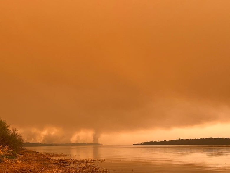 Wildfire smoke billowing into the air has created a large, thick smoke cloud covering the sky. The atmosphere has an orange tint. The cloud and smoke plumes hover over a large lake and some forest.