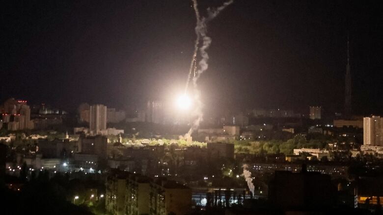 A large flash of light in the sky and a vapor trail appear over a city at night. 