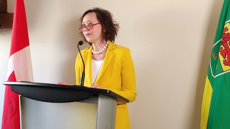 A white woman, with short brown air, is wearing glasses with a red frame, a white pearl necklace and a yellow suit with a white top underneath. She is standing at a podium with a microphone. A Canadian flag stands to her right.