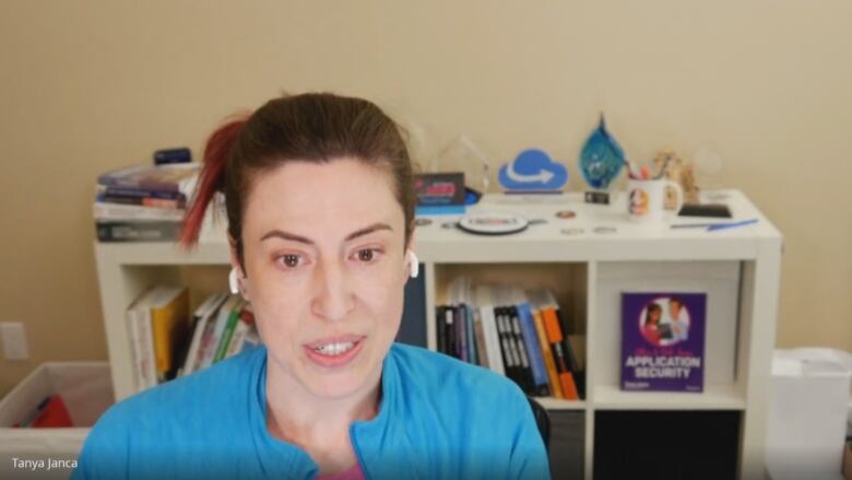 A woman with long dark hair pulled back from her face faces the camera. She is swearing a blue top and there is a bookshelf in the background.