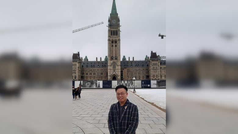 A black-haired man in a long jacket stands before Canada's looming Parliamentary tower.