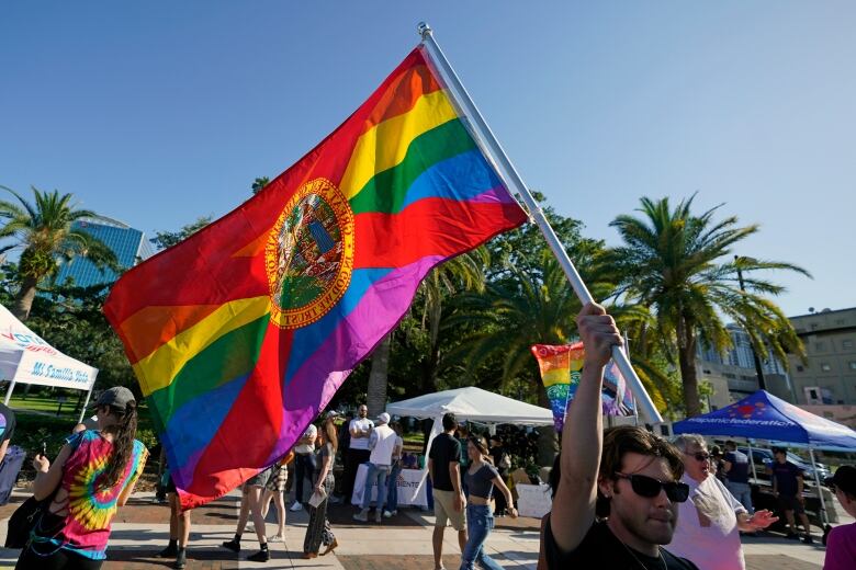 A man holds up a rainbow flag with a red lines crossing it and a state seal in the centre, while walking past people and palm trees in the background.