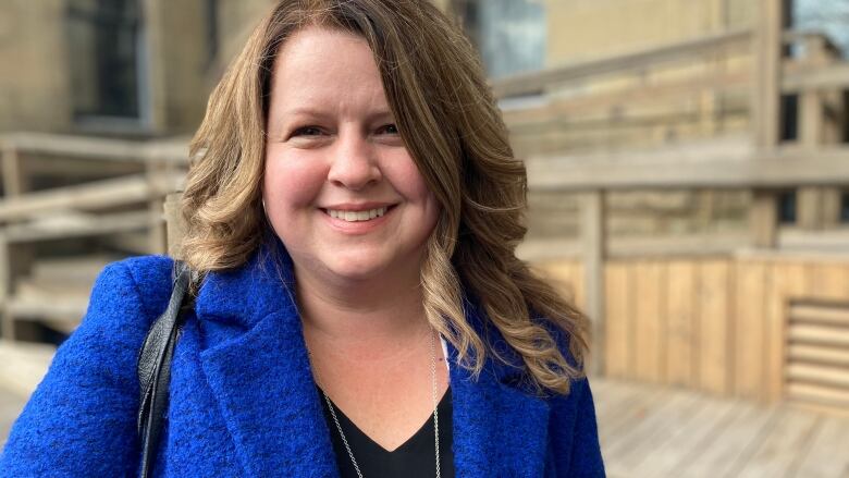 A woman with long light-brown hair, in a blue suit and black shirt stands outside the legislature and smiles for the camera.