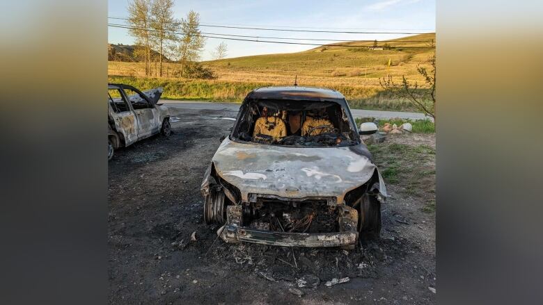 A burned white car, with a hill and utility cables as background.