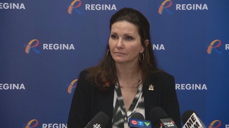 A middle-aged woman with long brown hair is wearing hoop earrings and dark blazer. There is a pin on her lapel. She is standing in front of a blue screen that features the word 'Regina' on it.