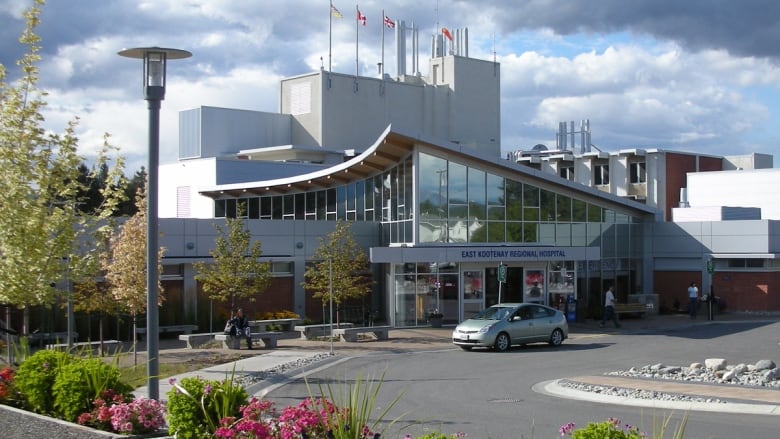 A silver car is parked outside the front doors of a grey hospital on a sunny day. A person is visible sitting at a picnic table.