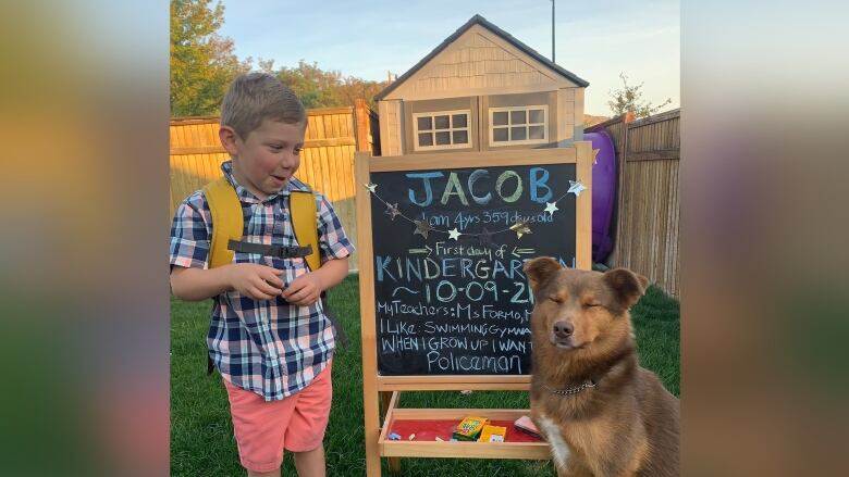 A boy in shirt and shorts is pictured with a backpack standing next to a dog in his backyard.