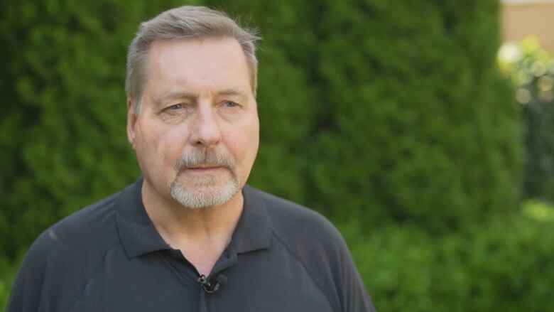 A man with short grey hair and blue eyes stands outside.