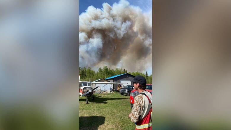 Plumes of smoke rise from behind a farmhouse, with a plane and vehicles visible in the background next to a white man.