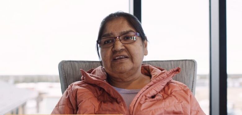 An Indigenous woman in a pink jacket sits in front of a window facing the camera