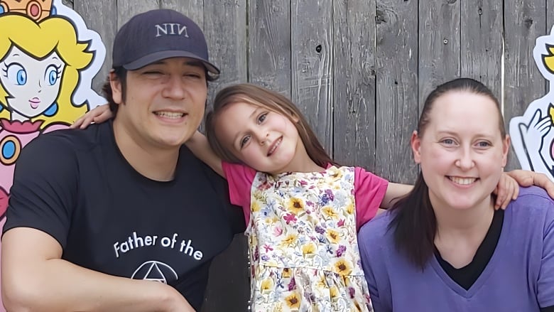 A mom and dad kneel down for a photo with their daughter, beaming at the camera. It is her birthday, a princess Peach themed birthday.