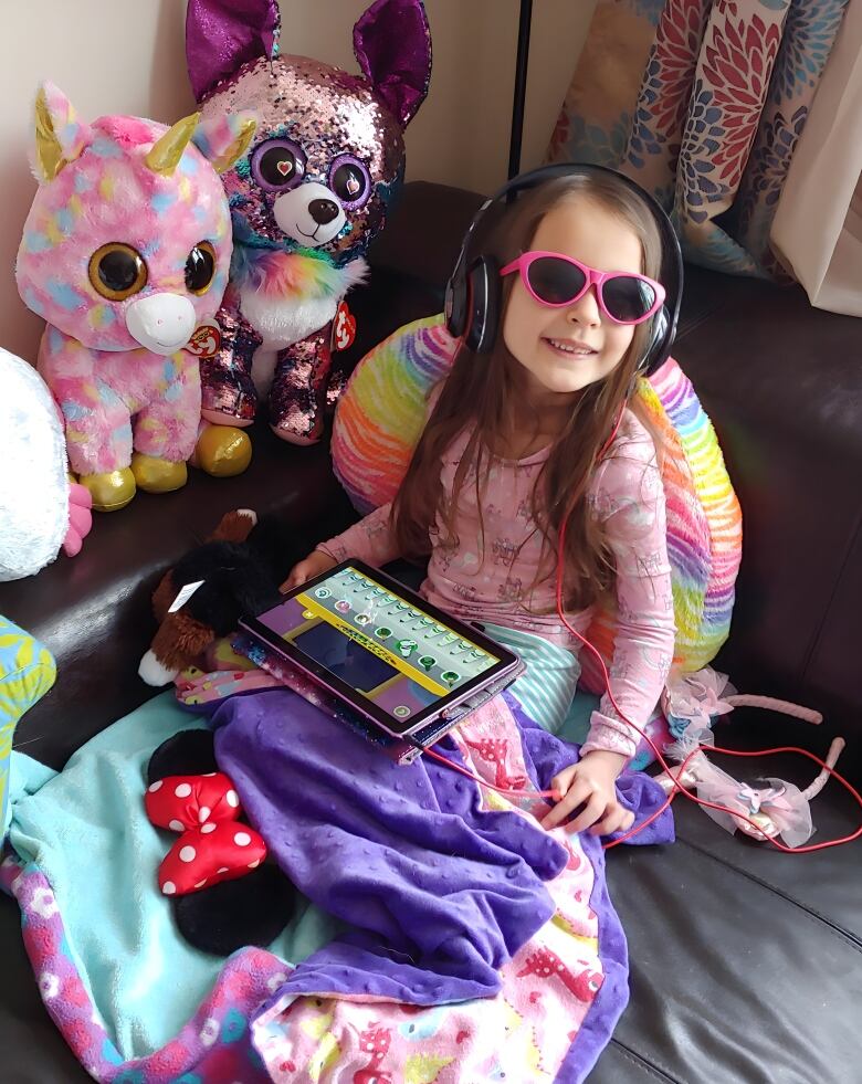 A young girl sits on a bed surrounded by stuffed animals. She wears headphones and sunglasses and has a tablet in her lap