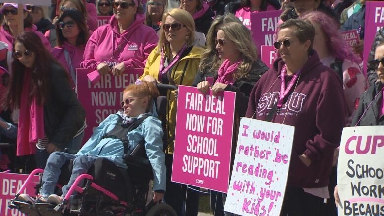 A crowd largely dressed in pink stand carrying signs that read 