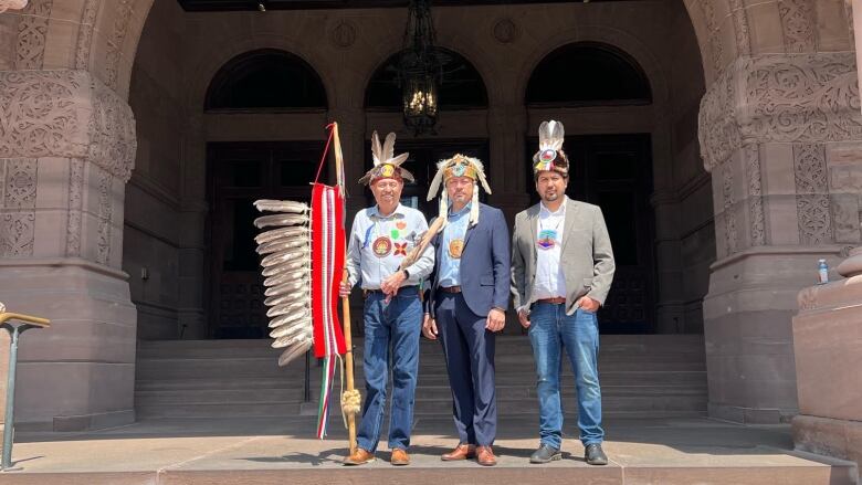 Three men wearing indigenous regalia standing under a stone arc.