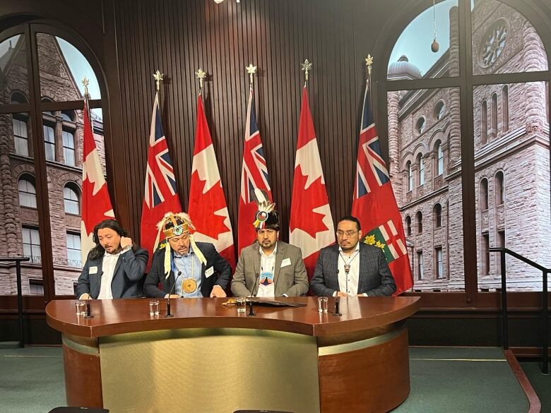 Four men sitting at a desk, with multiple flags behind them.