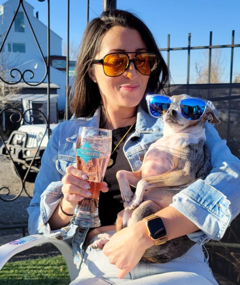 A woman holds a chihuahua in sunglasses while holding a beer.