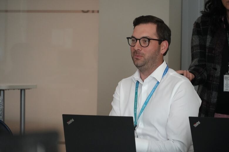 A man in a white shirt with a blue lanyard speaks at a table. 