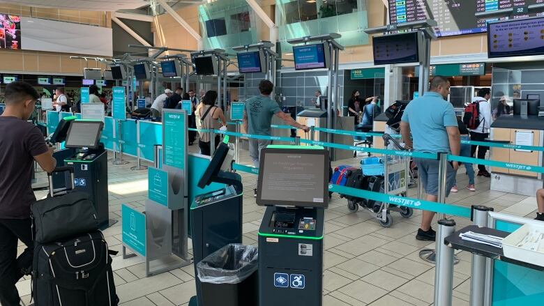 People line up at an airport counter in front of overhead monitors, some of which depict flight arrivals and departure times.