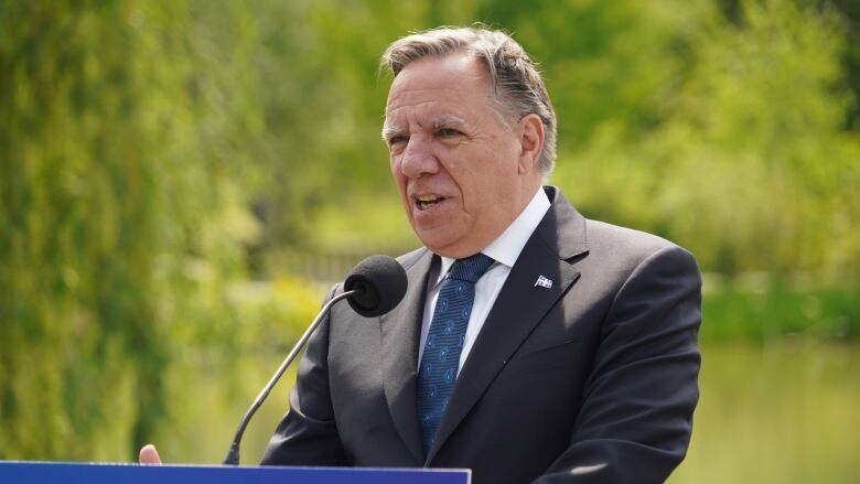 Franois Legault speaking behind a podium at an outdoor news conference. Greenery is seen in the background. 