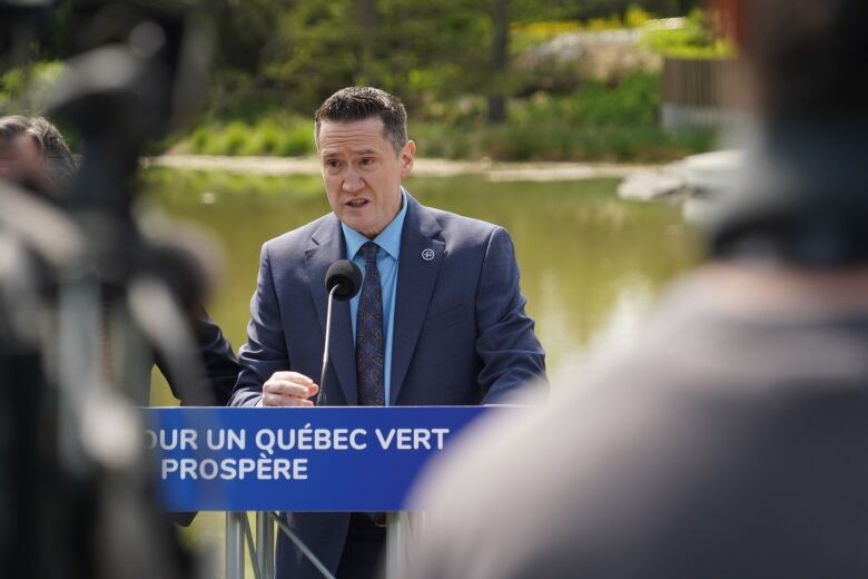 Benoit Charette speaking at a podium during a news conference outside. Greenery is in the background. Two cameramen are blurred in the foreground.