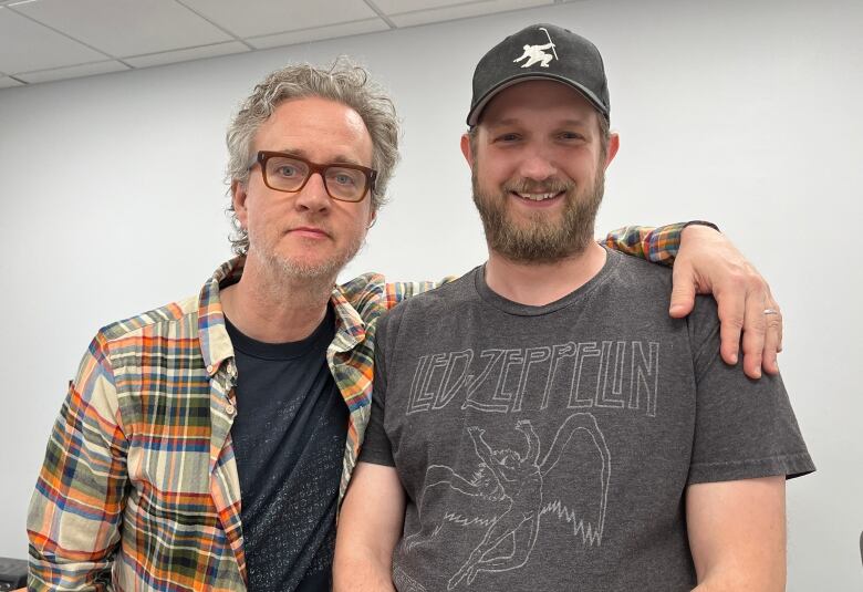 A man in a plaid shirt and black t-shirt stands on the left with his arm around a man on the right, wearing a hat and Led Zeppelin t-shirt. 