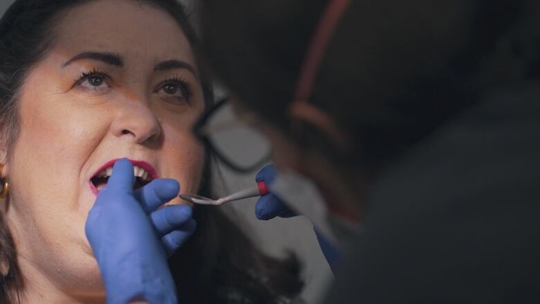 A white woman with dark hair and pink lipstick opens her mouth as the gloves hands of a dentist touch her teeth.