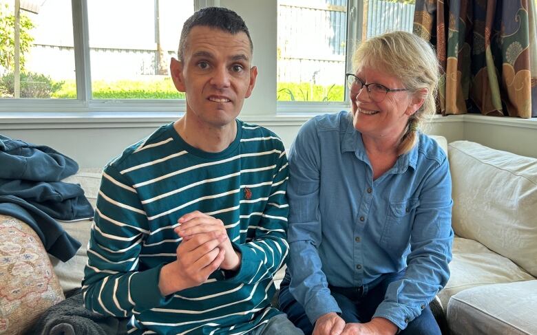 On the left, a white man with short salt and pepper hair, wearing a teal and white striped long-sleeved shirt, looks directly into the camera. He is sitting on a beige couch beside a blonde woman with glasses and a blue shirt who is smiling at him.