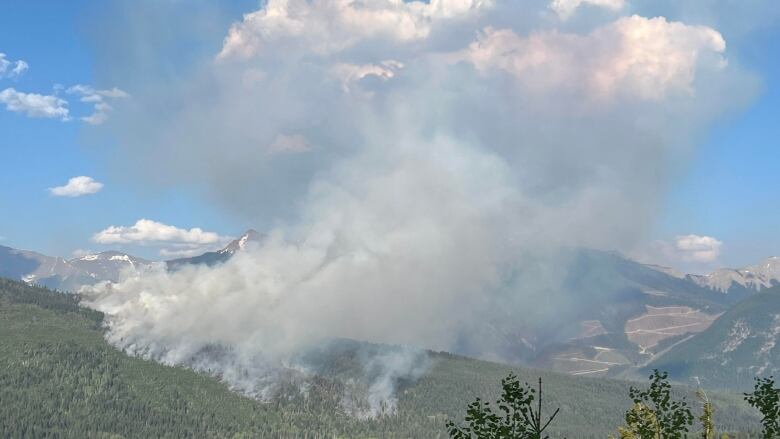 Smoke can be seen emerging from a heavily treed area. There are mountains in the background.