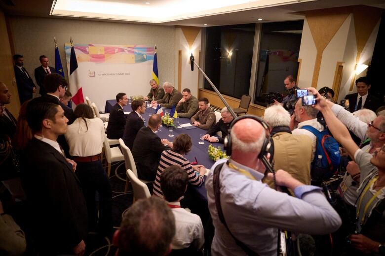 A group of people sit around a long table surrounded by people holding microphones and cameras.