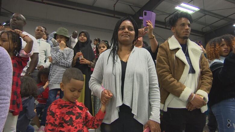 A crowd is pictured. In the centre, a woman holding her son's hand smiles and looks forward.