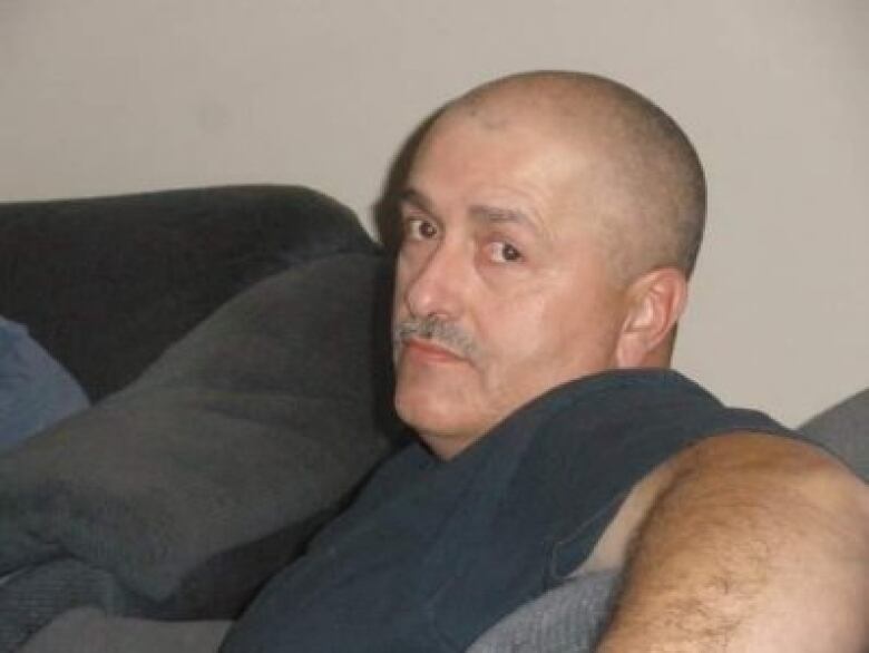 A man is seen sitting on a sofa and looking into the camera. He's wearing a black t-shirt.