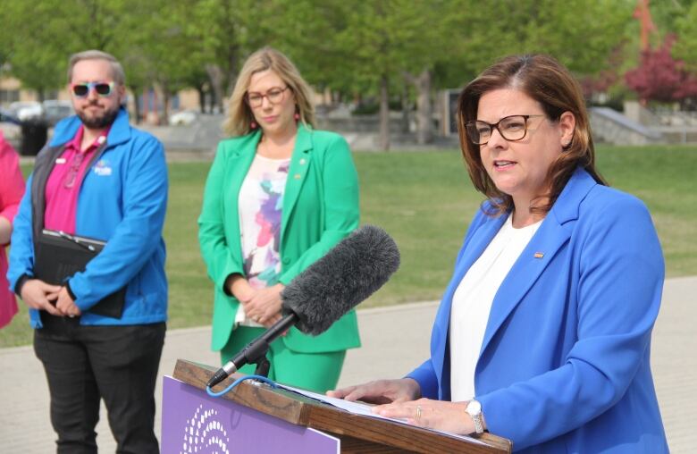 A woman in a blue blazer speaks behind the microphone at a news conference, while two other people stand behind them.