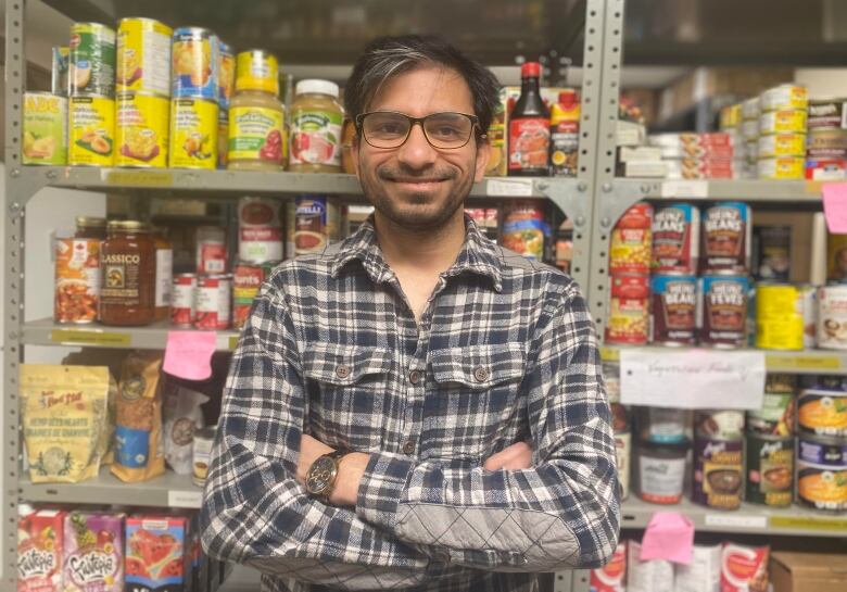 A man stands with his arms crossed, looking into the camera, with shelves filled with canned food behind him.