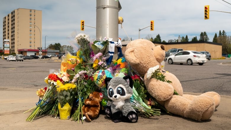 Flowers and stuffed animals are stacked by a lightpost on a sidewalk corner as a memorial. 