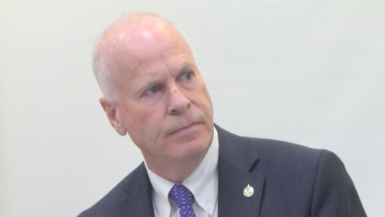 A man with short white hair looks to the right. He wears a grey blazer with a white shirt and a patterned tie with a pin on his lapel.