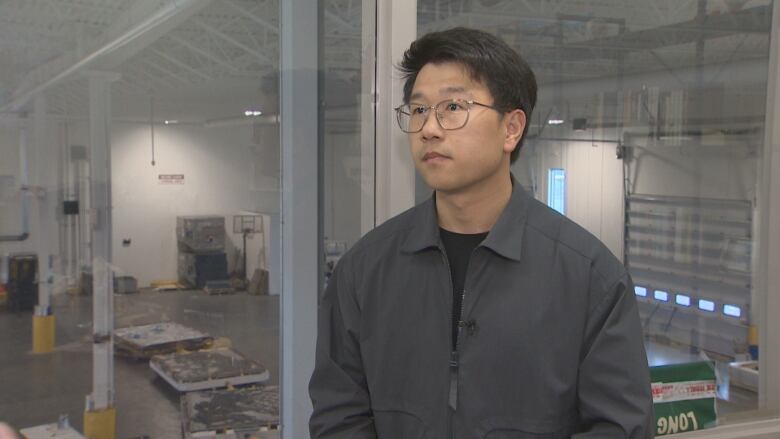 A man with dark hair and glasses wears a grey jacket with a black shirt. He is standing in a warehouse.