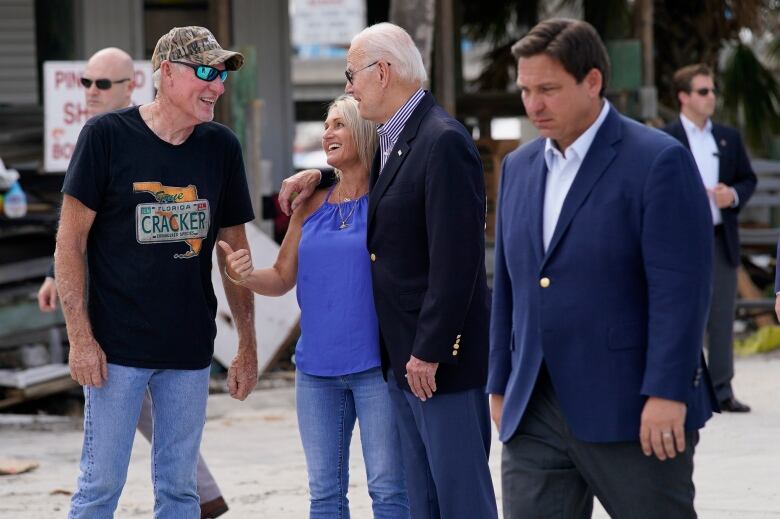 President Joe Biden, in aviators with a big smile, has his hand around a woman and is talking to a man. Ron DeSantis walks in the foreground, looking down.