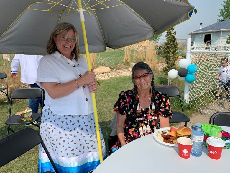 Mary Moonias sits at table while a second woman holds an umbrella to shield her from the sun.