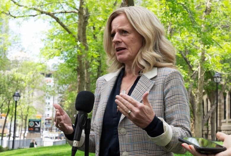 A woman speaks at a microphone outdoors, surrounded by trees.