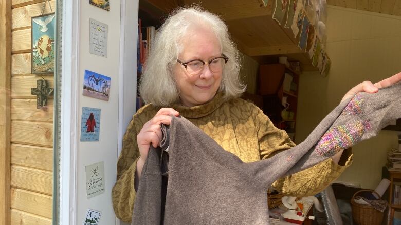 A woman stands in front of a wooden craft workshop, holding up a brown sweater with rainbow embroidered patches. 