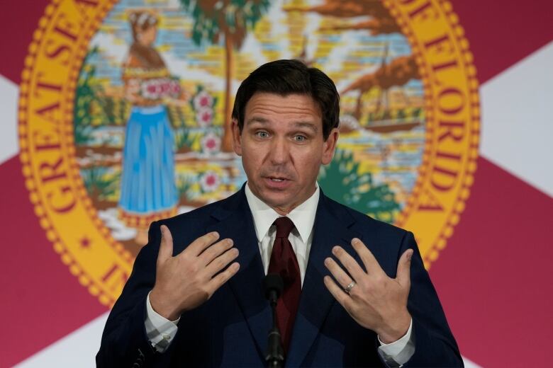 A person stands while making a speech in front of a large sign.