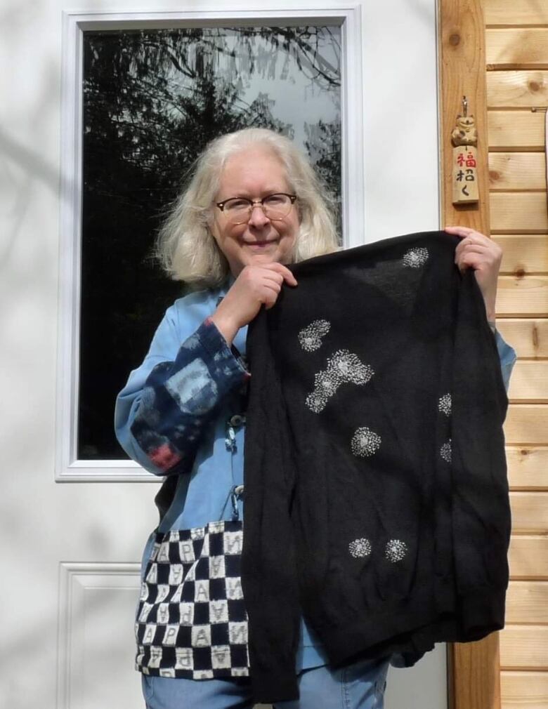 A woman is pictured holding a black sweater with white embroidery in clusters throughout the sweater. 