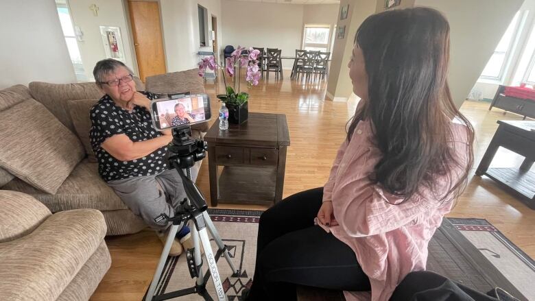 A young woman interviews an elder using a phone on a tripod.