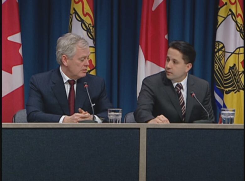 Two men in suits sit at a desk in front of the Canadian and New Brunswick flags.