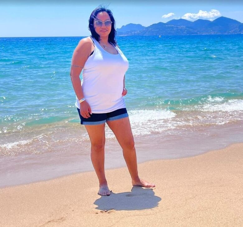 A woman in a white tank top and blue shorts stands on a sandy beach, with turquoise water, mountains and a light blue sky in the background.